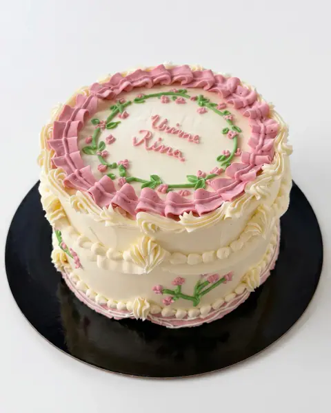 Cream vintage-style birthday cake with dusty pink ruffled piping, a piped floral wreath and hand-written lettering, a custom cake by Cakes By Vjollca in Pristina, Kosovo