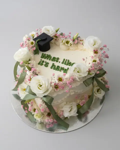 White heart-shaped graduation cake with a fondant graduation cap, fresh white lisianthus, pink wax flowers, sage ribbons and green lettering, a custom cake by Cakes By Vjollca in Pristina, Kosovo