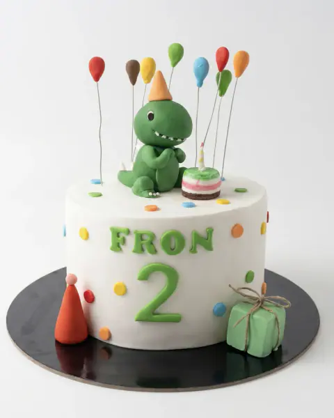 White birthday cake with a dinosaur figurine, balloons and confetti dots, a custom cake by Cakes By Vjollca in Pristina, Kosovo