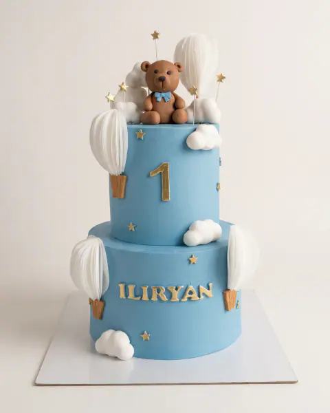 Blue two-tier first birthday cake with a fondant teddy bear, paper hot air balloons, clouds, gold stars and the child's name, a custom cake by Cakes By Vjollca in Pristina, Kosovo